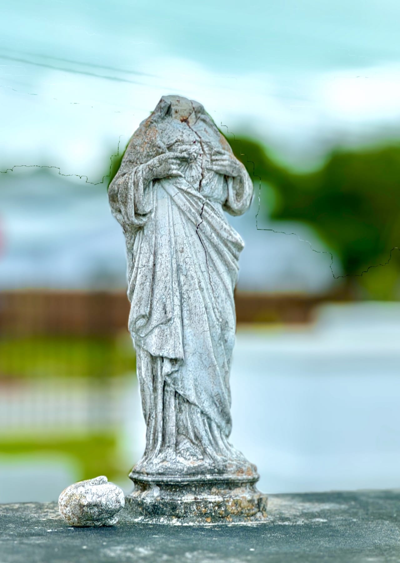 Color photograph of a cemetry statue depicting St. Francis. The background is blurred abstract blue and green. The statue's head has fallen off and is laying on the gorund.