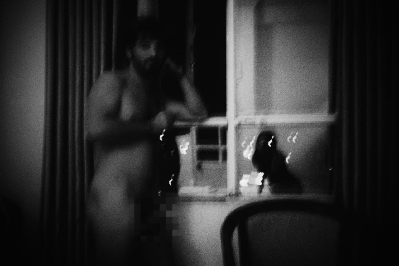 Dark and blurry black and white photo of a nude male (genitals obscured) in front of a draped window. The image is abstract.