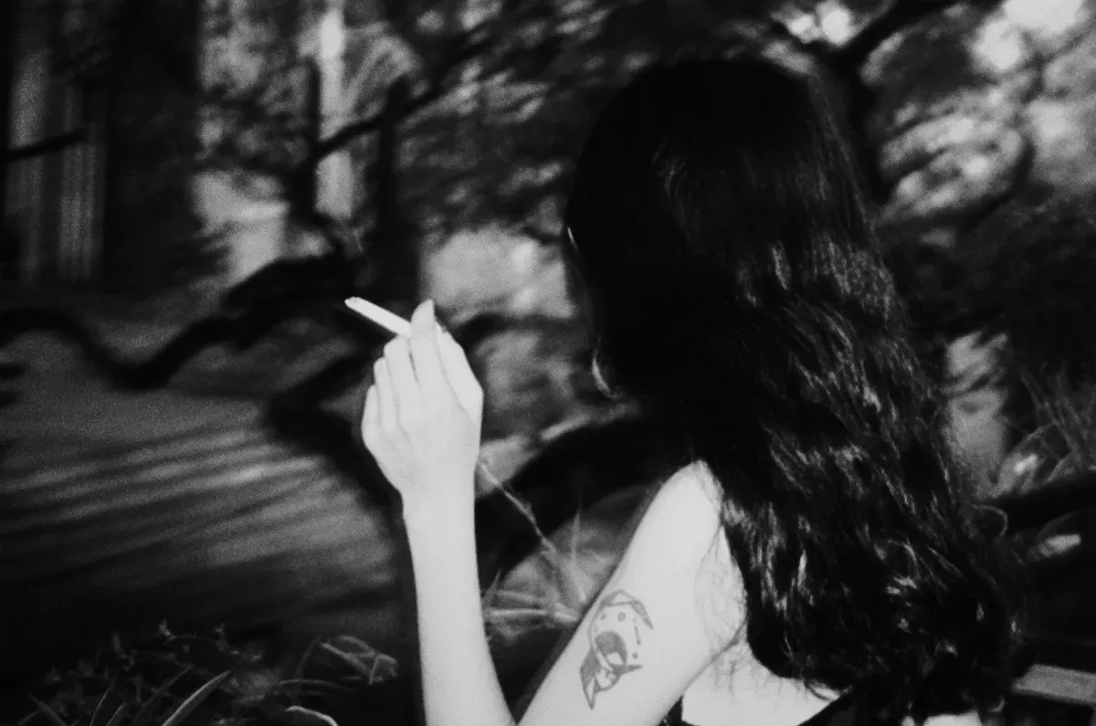 Black and white photo of the author with long dark hair, turned away form the camera. They are holding a cigarrette.