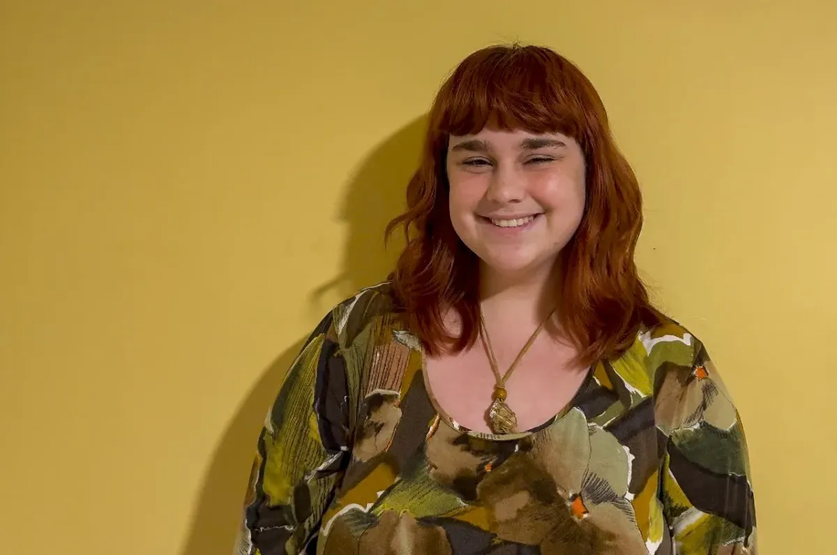 Carolina, with red hair, bangs and freckles is wearing a top with a floral pattern. She is smiling and looking at the camera