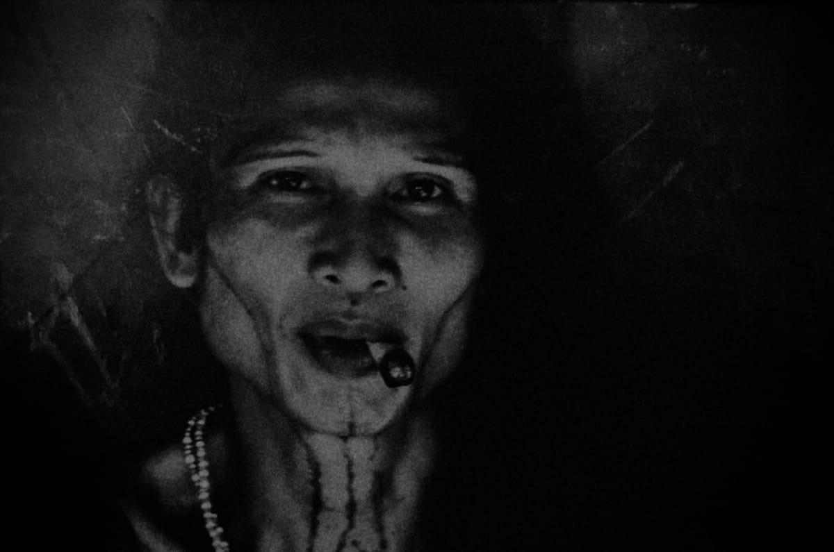 Very dark, grainy, low contrast photograph of an androgynous person with a cheroot in their mouth.