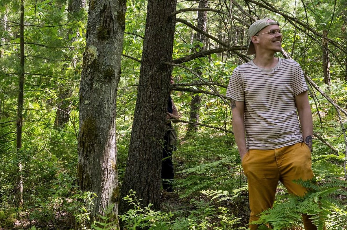 A guy standing in the woods with his hands in his pockets, looking away from the viewer. A figure peeks from behind a tree.