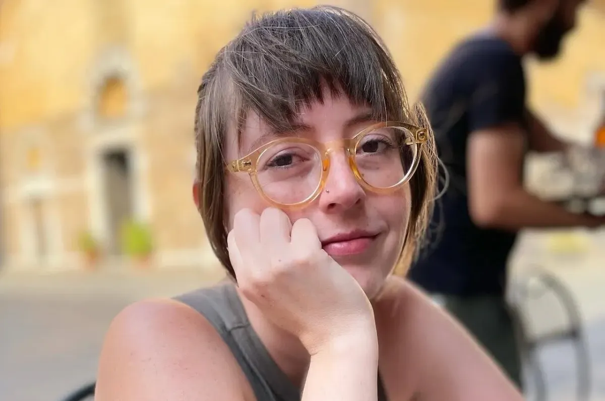 A white woman with freckles wearing yellow glasses. She has a small nose stud and her hair is brown with short tousled bangs