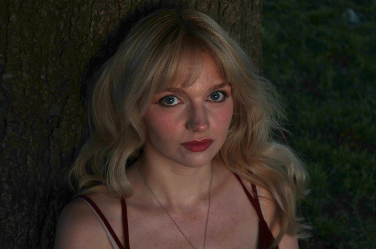 A woman with blue eyes and black eyeliner in a red summer dress seated, leaning against a tree. Her look is melancholy.