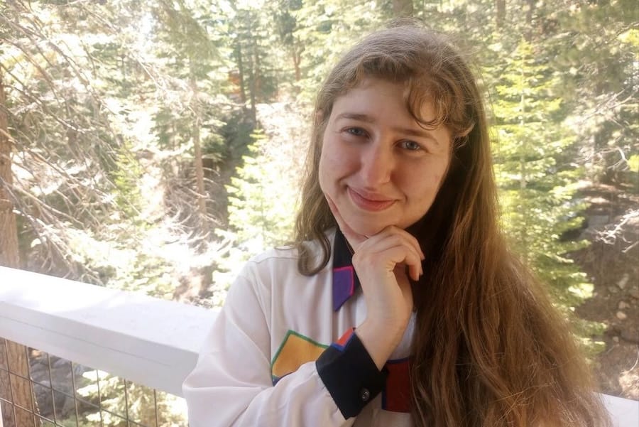 A white woman with long brown hair wearing a button down shirt with colorful squares on it, posing against a background of pine trees. She is smiling, her index finger alongside her chin.