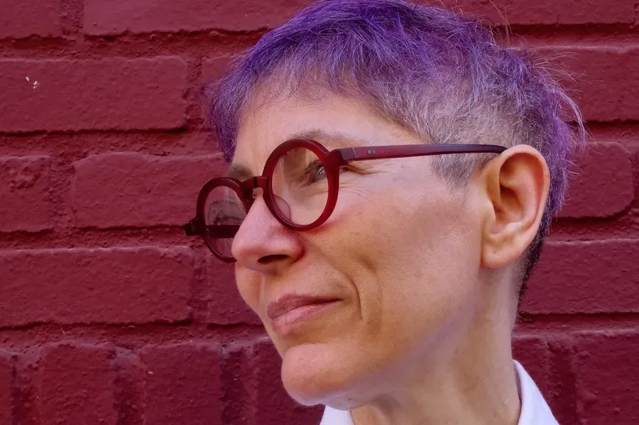Emma with short purple hair, red glasses, white shirt against red brick wall.