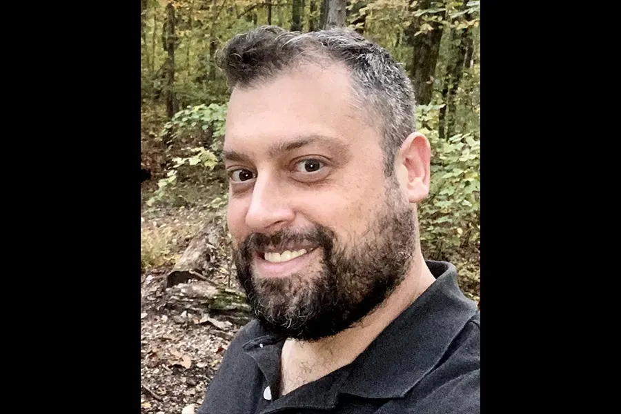 Poet Joshua St. Claire is standing in a forest. He is smiling with a brown beard.